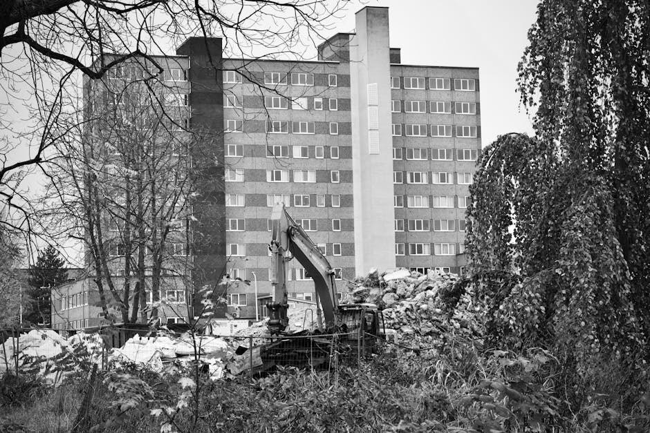 A construction excavator with a long arm and bucket attachment is positioned among a pile of rubble and debris in a garden area, surrounded by overgrown vegetation and leafless trees. The ground appea