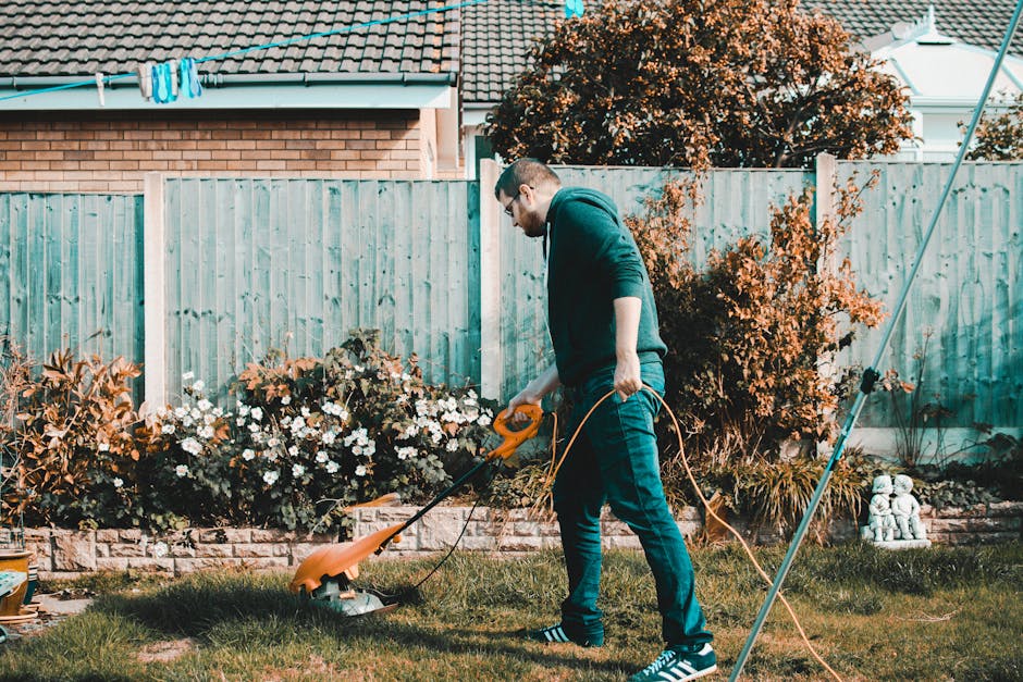 Close-up image showing a gardener's hands trimming a dense, well-maintained hedge with sharp, bright green garden shears. The gardener wears a red long-sleeve shirt, and the shears are positioned to r